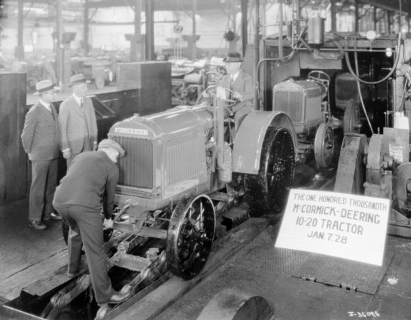 Men are standing around a tractor in a factory. A line man is tightening the last bolt. A sign next to the tractor reads: "The One Hundred Thousandth McCormick-Deering 10-20 tractor Jan. 7, '28."