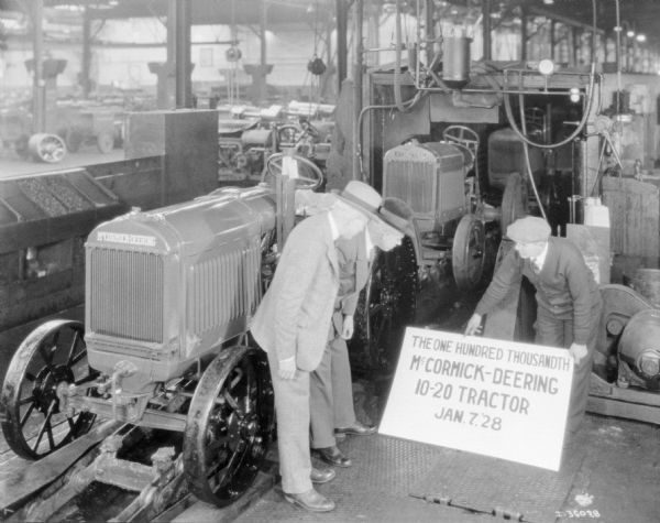 Men are standing near a tractor in a factory looking at a sign that reads: "The One Hundred Thousandth McCormick-Deering 10-20 tractor Jan. 7, '28."