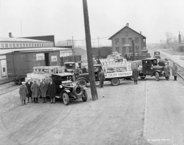 Elevated view of groups of men standing near trucks at a railroad station. Local IH dealers are standing by their trucks with banners on the side.