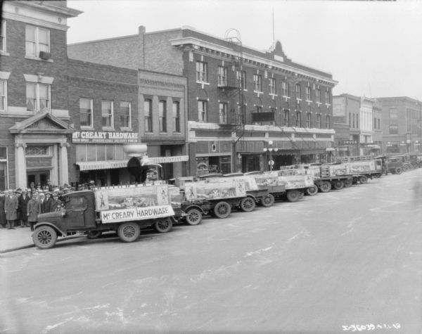 Elevated view across street towards a group of trucks from local IH dealers parked along the curb at an angle in front of storefronts. The McCreary hardware store is in the background. The trucks have banners on the side of their truck beds. There is also a large cream separator set up behind the trucks.