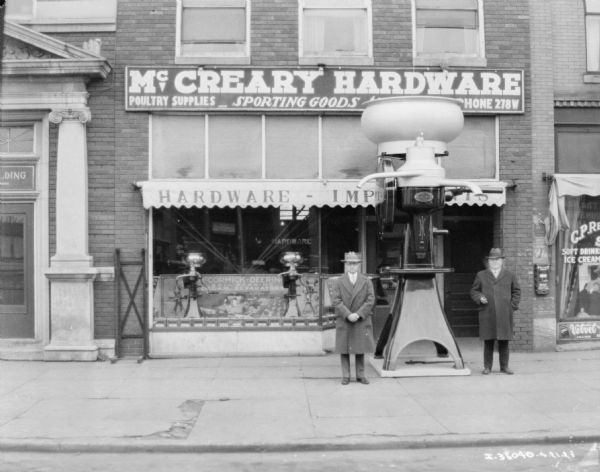 View from street towards two men standing on either side of a giant cream separator in front of the McCreary Hardware Store.