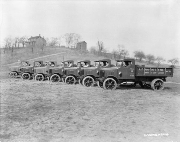 Seven men are sitting in the driver's seats of seven trucks parked in a clearing in a field. Behind them on a hill are buildings among trees. he sign painted on the truck at front right reads: "A.F. Jord and Co. Inc/. General Contractors."