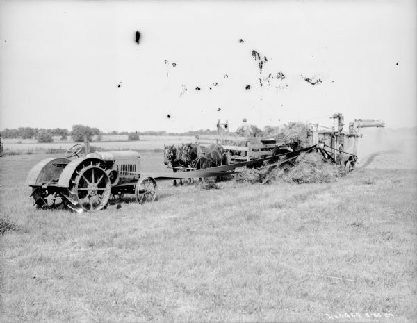 View across field towards a tractor in the left foreground. The tractor is belt-driving a hay press. In the center a man is standing on a wagon pulled by a team of horses.