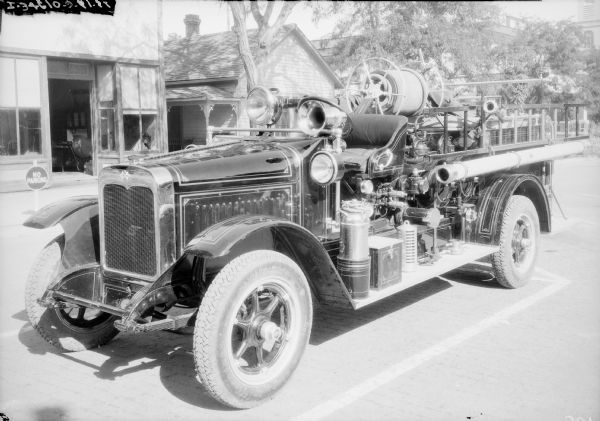 Three-quarter view from front left of a Model S-26 1927 fire truck parked in the street. In the background is a storefront.