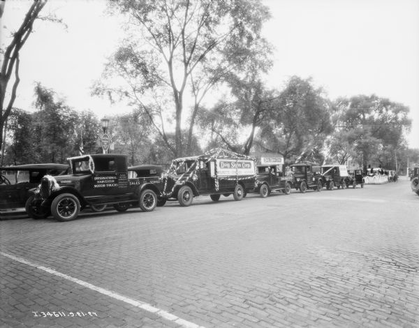 View across street toward a fleet of trucks lined up for a parade. An International Harvester Motor Trucks is decorated, and two other trucks have a sign for "Doig Signs Corp." A sign on a lamppost reads: "Welcome, Wilmette Day, Aug. 17th, 1927."