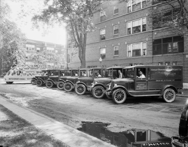 View towards a fleet of trucks for the Great Northern Laundry lined up for a parade. Men are sitting in the driver's seat of each truck which are parked at an angle along a curb. The truck at the back of the line is decorated, and has a sign at the top that reads: "Great Northern Laundry, Our Business is Clean," and a sign along the bottom that reads, in part: "International Moto___."[Presumably International Motor Trucks]