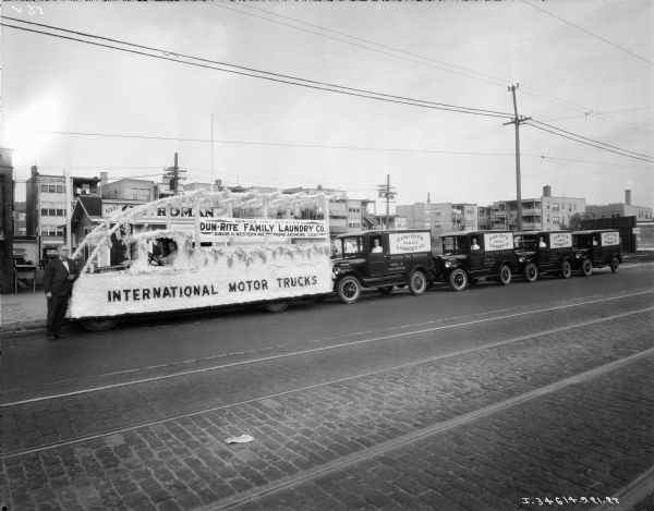 View from street towards a fleet of trucks parked along a sidewalk. The truck on the far left is decorated for a parade, and a sign on top of the truck reads: "Dun-Rite Family Laundry Co."