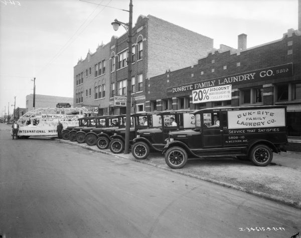 View from street towards a fleet of trucks lined up along a sidewalk at an angle. The truck at the end is decorated for a parade, and a sign on top of the truck, and on the building behind the fleet, reads: "Dun-Rite Family Laundry Co."