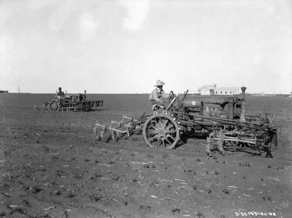 View across field towards two men driving tractors across a field pulling cultivators. The right side of the tractor has a sign for "IHC" over the painted "Farmall" sign. Farm buildings are along the horizon in the background.