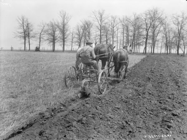 Three-quarter view from right rear of a man on a horse-drawn plow working in a field. Three horses are pulling the man and plow. In the background an automobile is parked under trees near a fence.