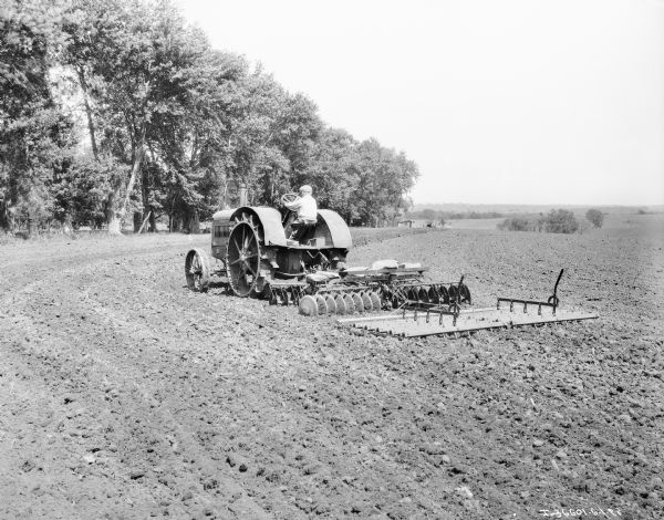 Three-quarter rear view from left of a person (may be a child) driving a 15-30 tractor pulling an implement in a field. Trees line the edge of the field in the background on the left.