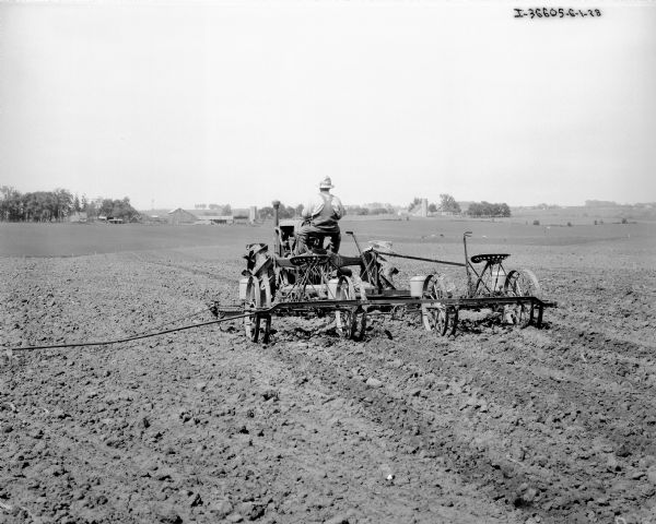 Rear view of a man driving a Farmall tractor pulling a planter in a field. Farm buildings are in the far background.