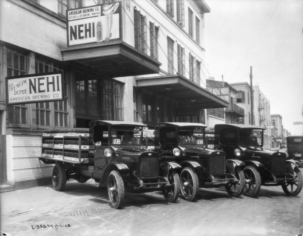 Three delivery trucks are backed up to the loading dock of the American Brewing Co. The back of the trucks are loaded with crates of Nehi bottles.