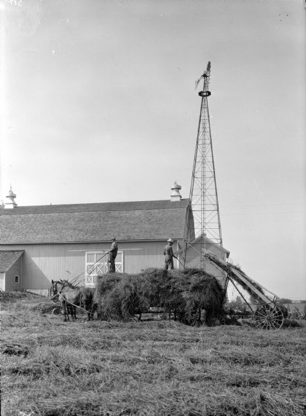 View across field towards a man and a boy standing on hay on top of a horse-drawn wagon. On the right is a hay loader filling the wagon. In the background is a large barn and a windmill.
