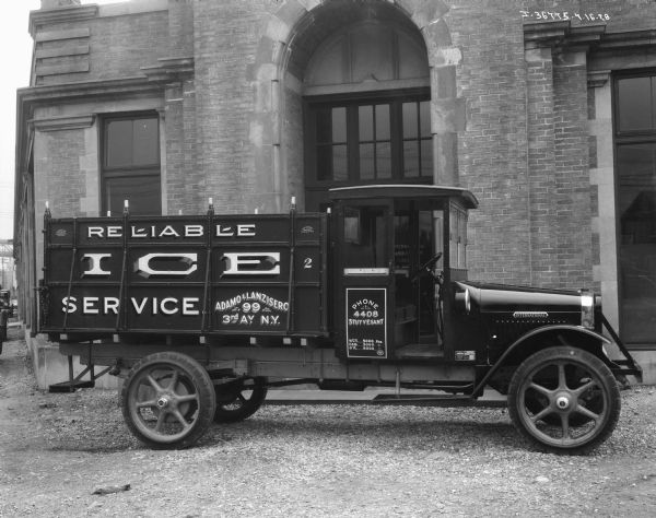 Right side view of a International truck used by the Reliable Ice Service, Adamo & Lanzisero, for ice delivery parked in front of a brick and stone building with an arched entrance. The sign on the glass door of the building reads: "International Harvester Company of America."