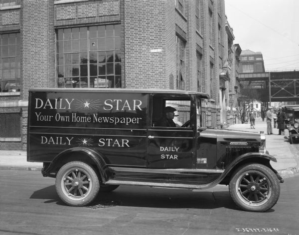 View across street towards a man driving a Daily Start "Your Own Home Newspaper" delivery truck near a city block.