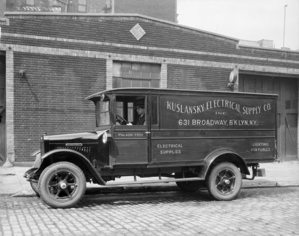 View towards a man sitting in the driver's seat of an International truck parked along a curb in front of a brick industrial building.