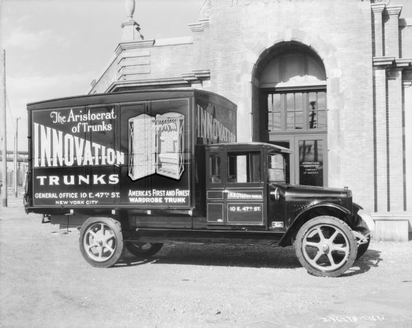 Right side view of a truck parked in front of the arched entrance of a stone and brick building with an arched entrance. The sign on the glass door of the building reads: "International Harvester Company of America." The sign on the truck reads: "The Aristocrat of Trunks, Innovation (Trademark) Trunks, America's First and finest Wardrobe Trunk."
