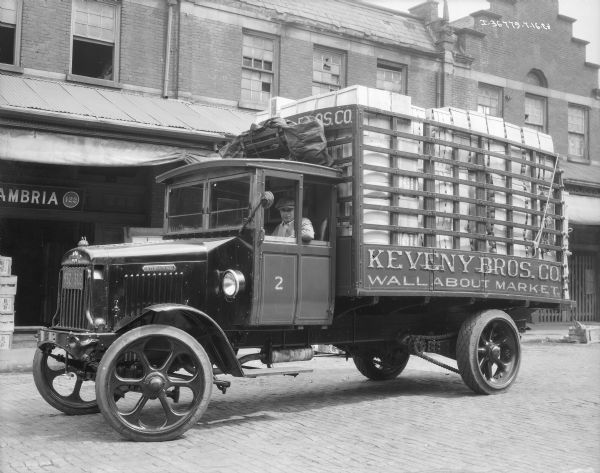 View across cobblestone street towards a man sitting in the driver's seat of a delivery truck for Keveny Bros. Co. Wallabout Market. The truck has a stake body, and there are crates piled high to the top. On the front of the truck is a Commercial NY 28 license plate. Industrial brick buildings are in the background.