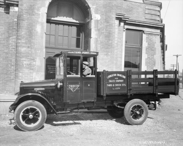 View of a man sitting in the driver's seat of a truck parked in front of a brick and stone building with an arched entrance. The sign on the glass door of the building reads: "International Harvester Company of America." 