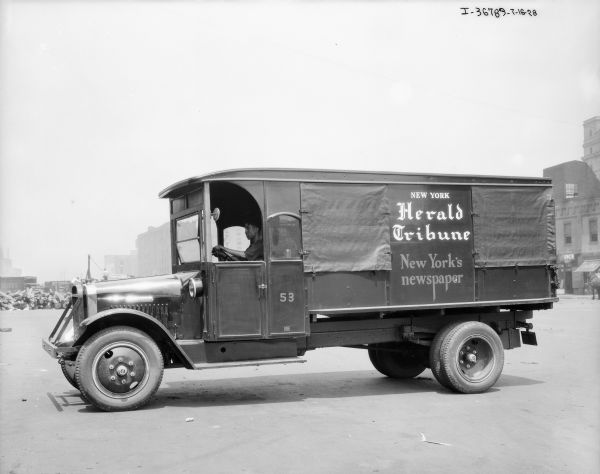 View towards a man sitting in the driver's seat of a truck. Buildings are in the far background and on the right. The sign on the truck reads: "New York Herald Tribune, New York's newspaper."