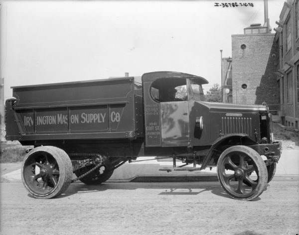 View towards the passenger side of a truck. A man is sitting in the driver's seat. The sign painted on the side of the high truck bed reads: "Irvington Mason Supply Co." An industrial brick building is on the right.