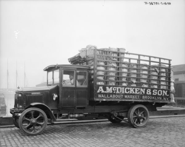View across street towards a man sitting in the driver's seat of a truck for A. McDicken & Son delivery truck. The truck bed has a stake body, and is stacked high with wood barrels and round wood crates carrying vegetables.
