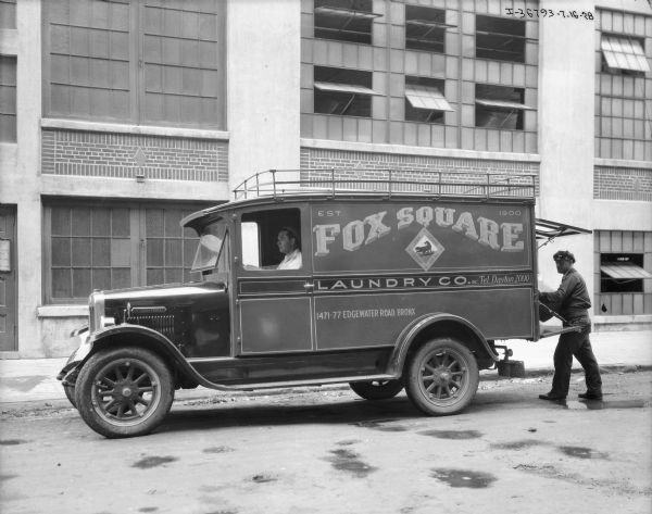 View across street towards a man sitting in the driver's seat of a Fox Square Laundry Co. truck. A man wearing a hat and uniform is standing at the open back of the truck, either loading or unloading. In the background is an industrial building.