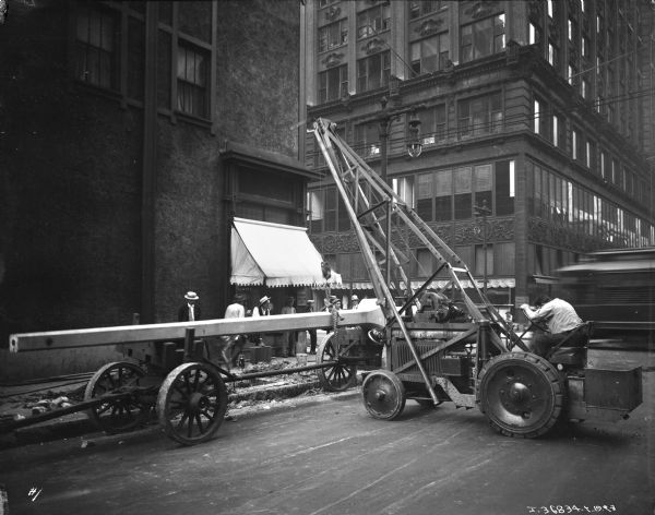 View across street towards a man using a hoist to mount street lights a near an intersection in a city. Men are standing on the sidewalk watching the installation. A group of men in work clothes are helping to set the light in place. Commercial buildings are in the background, and on the right is the blurred image of a streetcar. 