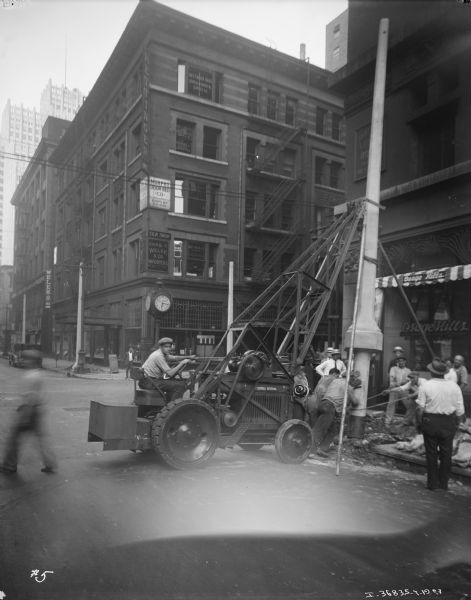 View across street towards a man using a hoist to mount street lights a near an intersection in a city. Men are standing on the sidewalk watching the installation. A group of men in work clothes are helping to set the light in place. Commercial buildings are in the background.