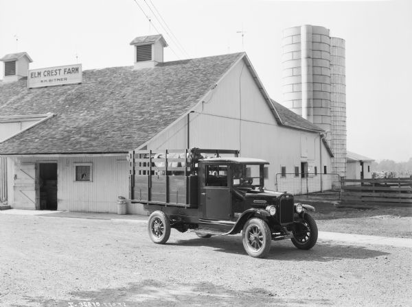 View across farmyard towards a man sitting in the driver's seat of an International truck. Two cows are standing in the back of the truck which has a stake body. The sign on the barn in the background reads: "Elm Crest Farm, W.H. Bitner." An Ohio license plate is on the front of the truck which has Goodyear tires. On the front above the grill is the International triple diamond logo. 
