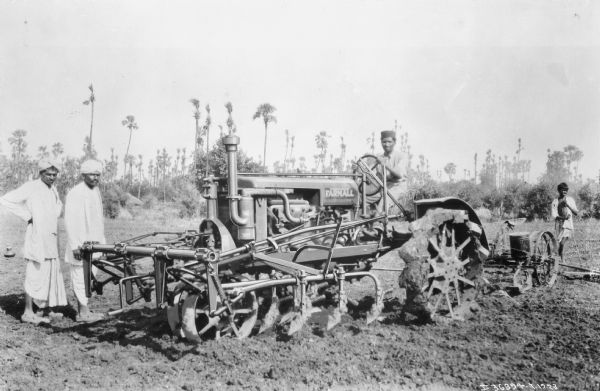 A man is sitting on a Farmall tractor with a cultivator in a field. Two men are standing near the tractor on the left, and another man is standing behind the tractor on the right.