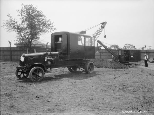 An International truck with a tractor fitted with a crane in a cab on the back. Sign on the side of the truck reads: "Truckrane." A sign on the base of the truck bed reads: "Hanson." Men are standing near a pile of dirt behind the crane on the right. There is a tall, wood fence in the background.