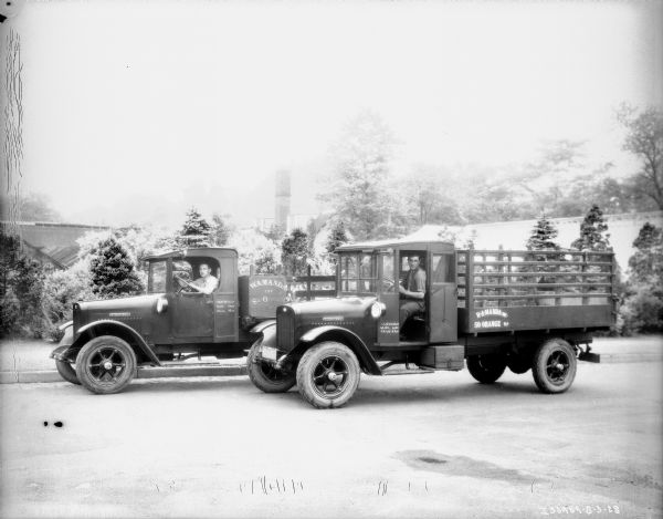 View towards two men, each sitting in the driver's seat of a delivery truck parked outdoors. Both trucks have stake bodies.