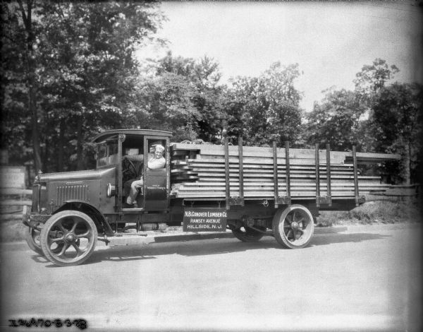 View across street towards a man sitting in the driver's seat of a lumber delivery truck. The sign on the side of the truck bed reads: "N.B. Conover Lumber Co., Ramsey Avenue, Hillside, N.J." The open truck bed is loaded with lumber.