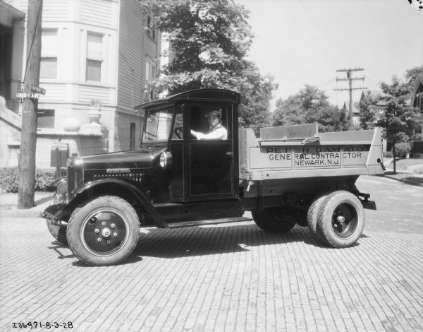 View towards a man sitting in the driver's seat of an International truck parked on a cobblestone street. The street signs on a power pole in the background read: "Bryant St." and "Lincoln Ave." The sign painted on the truck bed reads: "Peter D'Amato, General Contractor, Newark, N.J."