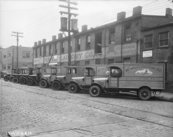 View across cobblestone street towards a fleet of about nine trucks parked at an angle against the opposite curb. Behind them is a long brick industrial building with a sign painted above the first-story windows that reads: "Home of Perfect Laundry Co." The signs painted on the side of the trucks reads: "We Wash Everything but the" which is over an illustration of a baby laying on a blanket.