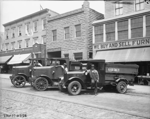 View across street towards three men with coal delivery trucks parked in front of storefronts. Two of the men are standing near the driver's side door, and the man at the end is sitting in the driver's seat of a truck. The sign on the trucks, and on a brick building in the background read: "R.H. See Coal Co."