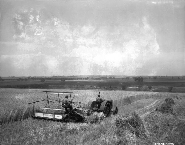 Slightly elevated view of a man driving a McCormick-Deering 15-30 tractor pulling another man on a McCormick-Deering binder in a field. Fields and farm buildings are in the distance.