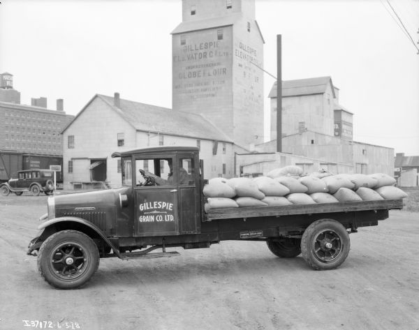 View towards a man sitting in the driver's seat of a Gillespie Grain Co. Ltd. truck, with an open truck bed loaded with sacks of oats. In the background is the Gillespie Grain Co. elevator, and other buildings.