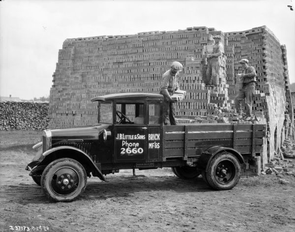 View towards three men loading a truck with bricks from a large pile. The sign painted on the truck reads: "J.B. Little & Sons, Brick Mf'gs."