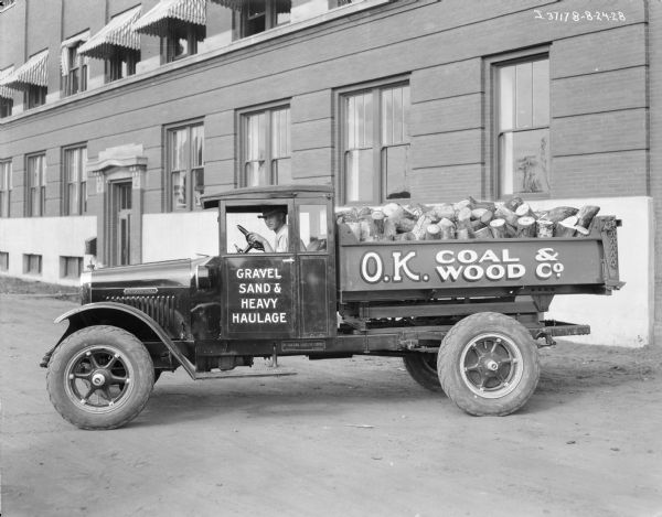 A man is sitting in the driver's seat of a truck loaded with firewood. The signs painted on the truck read: "Gravel Sand & Heavy Haulage: and O.K. Coal & Wood Co. A large brick building with striped awnings over some of the windows is just behind the truck.
