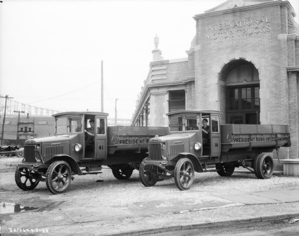 Two men are sitting in the driver's seats of two trucks. They are parked in front of a brick building with an arched entrance. The sign above the arch reads: "International Motor Trucks." There is a bridge is in the background on the left.