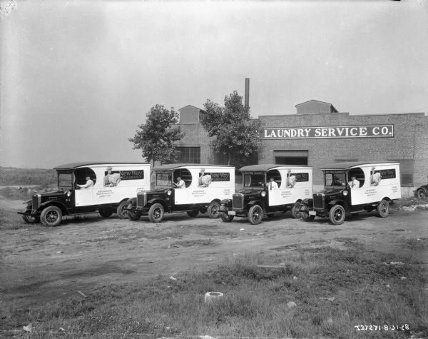 View towards four men sitting in the driver's seats of four laundry trucks. Behind them is a brick building with a sign for "[obscured] Laundry Service Co."