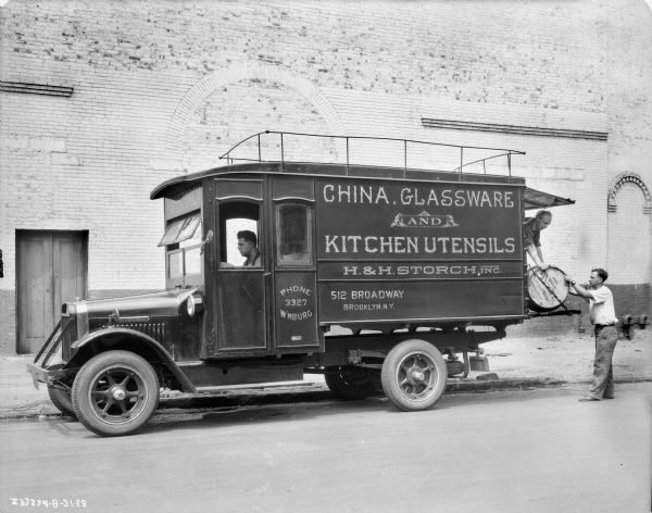 View across street towards a man sitting in the driver's seat of a truck parked along a curb in front of a brick building. Two me are loading or unloading a barrel at the back of the truck. The sign painted on the side of the truck reads: "China, Glassware and Kitchen Utensils, H. & H. Storch, Inc."