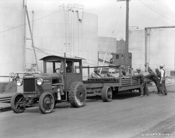 A man is sitting in the cab of an industrial tractor, while men are unloading pipes from the cart/trailer hitched to the tractor. Storage tanks are in the background.
