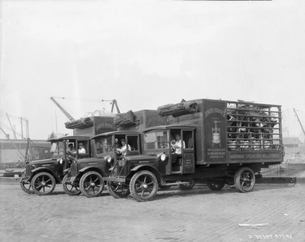 Three delivery trucks are parked in a row on a waterfront. Two to three men are sitting in each cab, and the signs painted on the trucks reads: "Louis Birner, Hardware Specialties, Woodenware." Each truck has a large load in the high-sided truck beds, and canvas is rolled in a wood shelf above each cab roof.