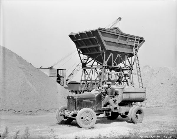 View of a man sitting in the driver's seat of an open truck being loaded by a large hopper for material for laying asphalt. Behind the hopper is a crane, which is partially obscured by a large pile of material.