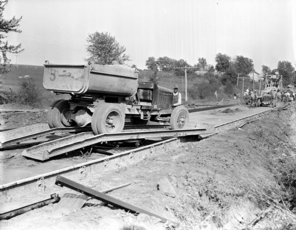 A man is standing at the front of a truck parked on a ramp. Further back on the right a group of men are working laying asphalt with large machinery. A group of people are watching them work in the background.