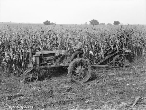 Left side view of a man driving a Farmall tractor pulling a corn picker in a cornfield.